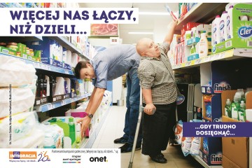 Dwóch mężczyzn przy sklepowych półkach. Wysoki mężczyzna nie może sięgnąć produktów z dolnych półek, a niskorosły z górnych. Przed nimi napis: Więcej nas łączy, niż dzieli